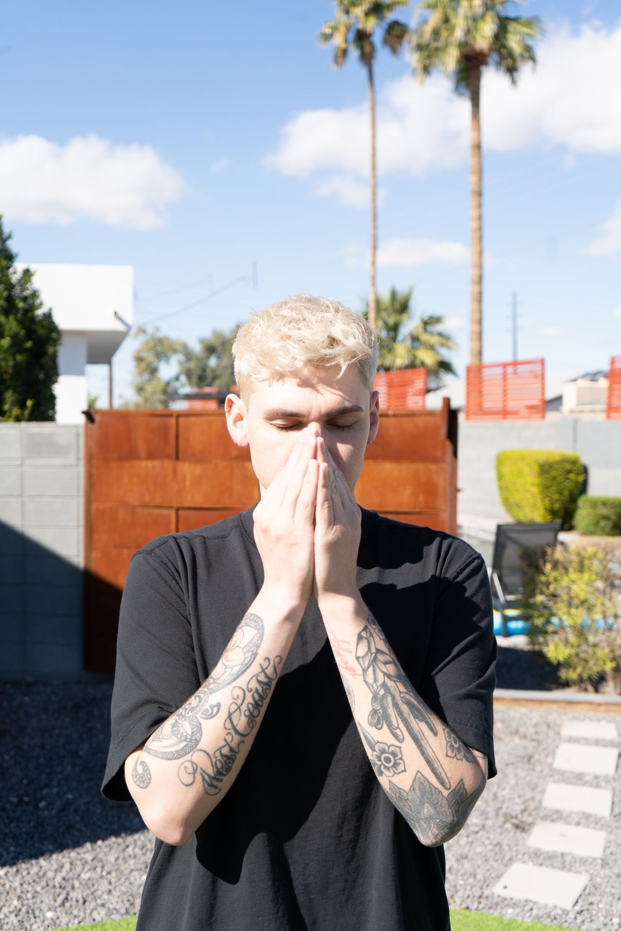 Person with tattoos covering their face outdoors on a sunny day, with palm trees in the background.