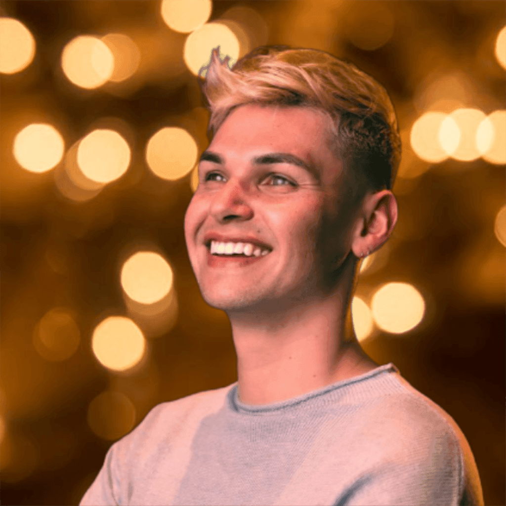 Smiling person with blond hair in front of a background with blurred golden lights.